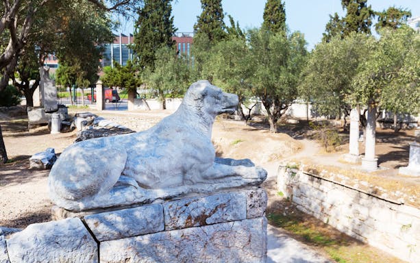 Stone dog statue at Kerameikos archaeological site, Athens, surrounded by trees and ancient ruins.