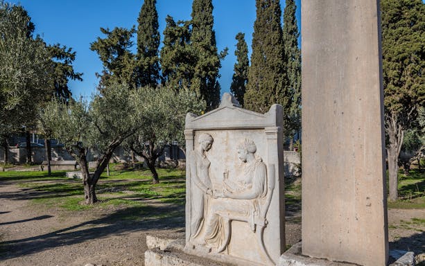 Ancient gravestone relief in Kerameikos, Athens, surrounded by olive trees.