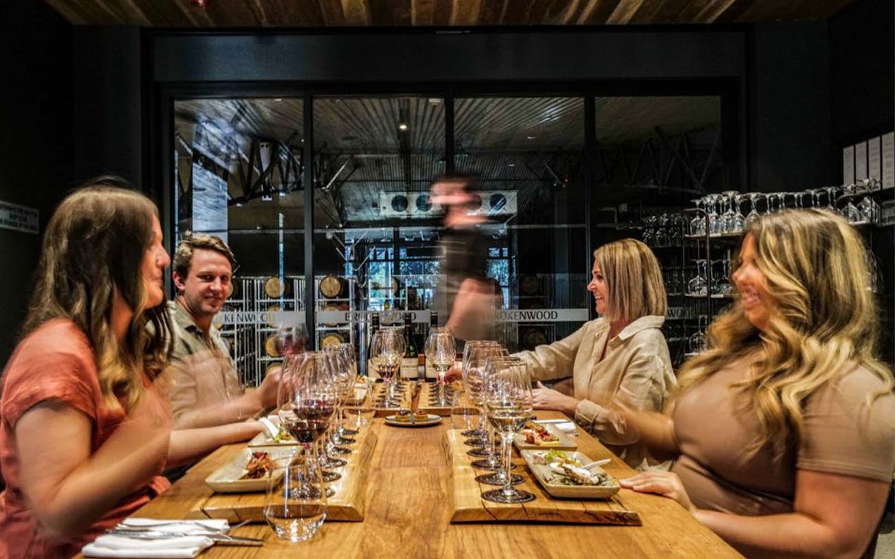 Guests enjoying wine tasting at Brokenwood winery, seated at a long table with wine glasses and plates.