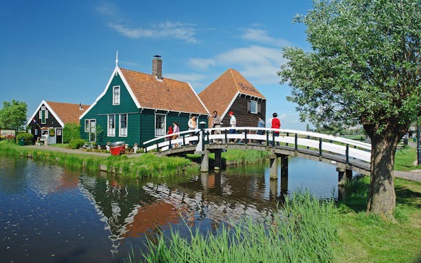 Visitors crossing a wooden bridge at Zaanse Schans, traditional Dutch houses in the background.