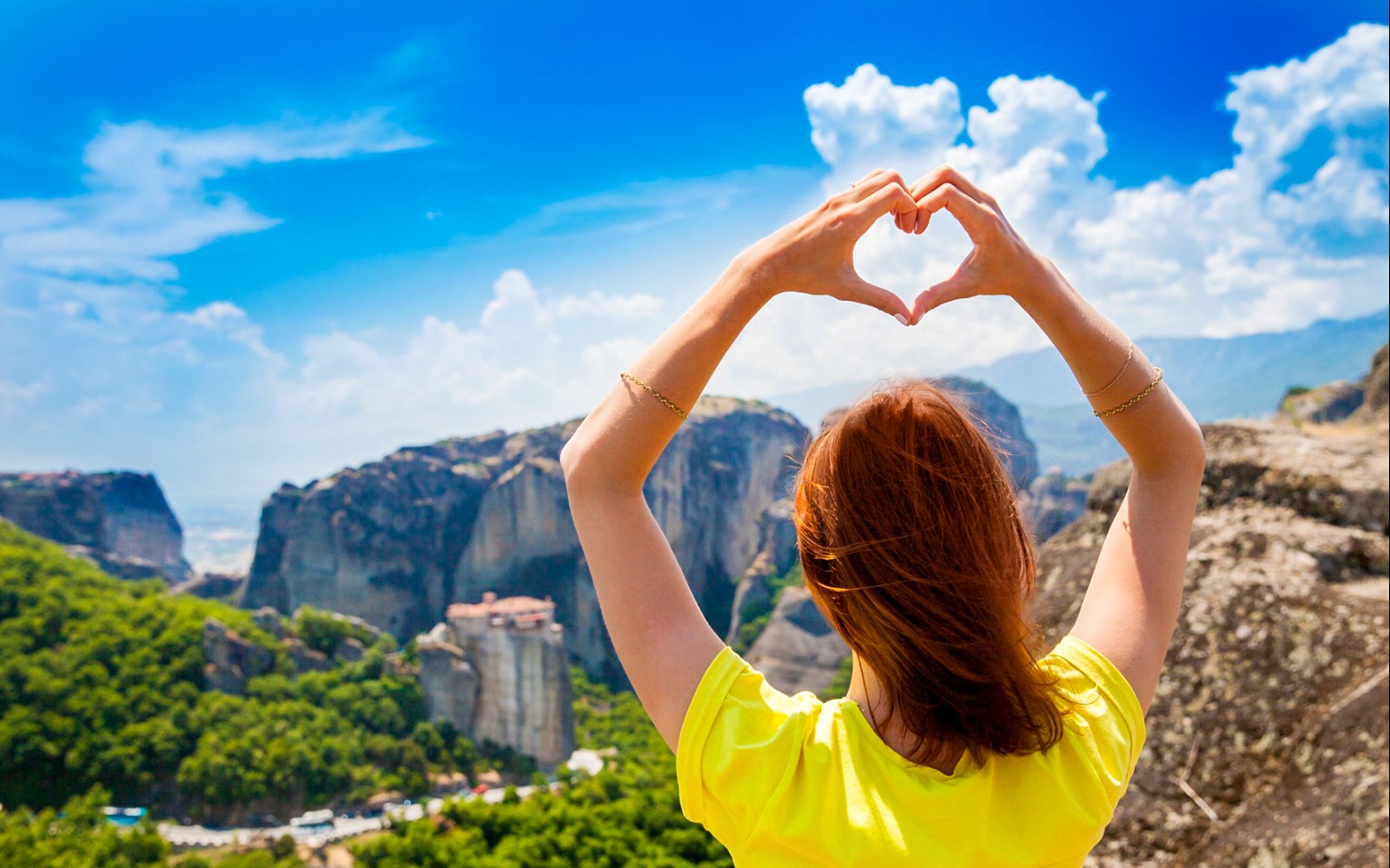 Person forming heart shape with hands overlooking Meteora rock formations.