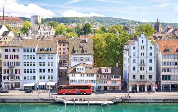 Historic buildings along the Limmat River in Zurich, with a tour bus parked nearby.