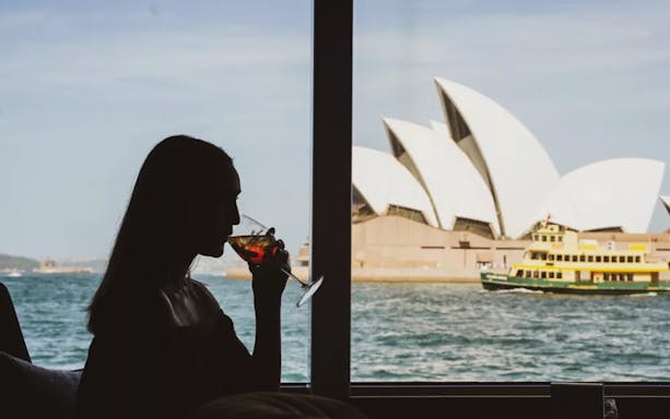 Person enjoying a drink with a view of the Sydney Opera House and ferry on the water.