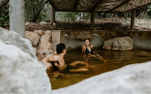 Couple relaxing in a natural hot spring pool at Peninsula Hot Springs, surrounded by rocks and greenery.