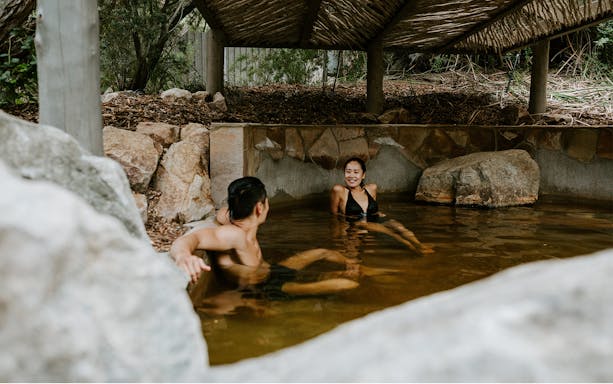 Couple relaxing in a natural hot spring pool at Peninsula Hot Springs, surrounded by rocks and greenery.