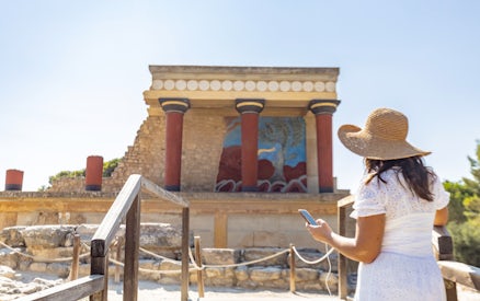 Visitor using phone for audio tour at Knossos Palace, Crete, with ancient fresco and columns.