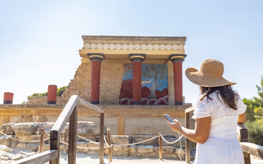 Visitor using phone for audio tour at Knossos Palace, Crete, with ancient fresco and columns.
