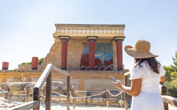 Visitor using phone for audio tour at Knossos Palace, Crete, with ancient fresco and columns.