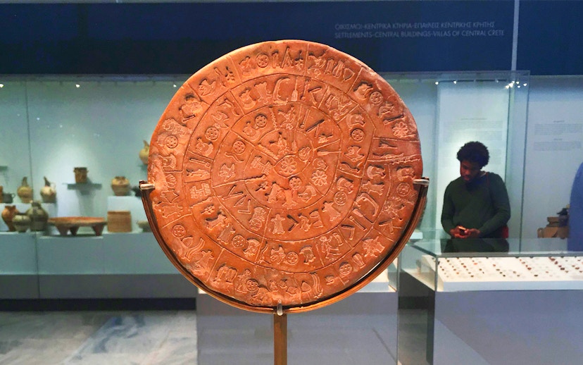Phaistos Disc at Heraklion Archaeological Museum, Crete, with visitors in the background.