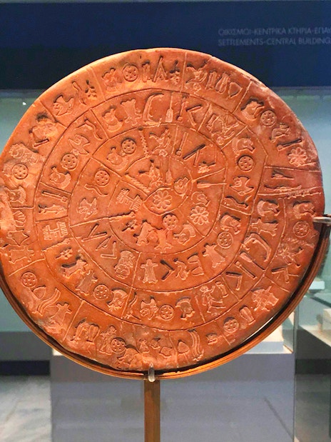 Phaistos Disc at Heraklion Archaeological Museum, Crete, with visitors in the background.