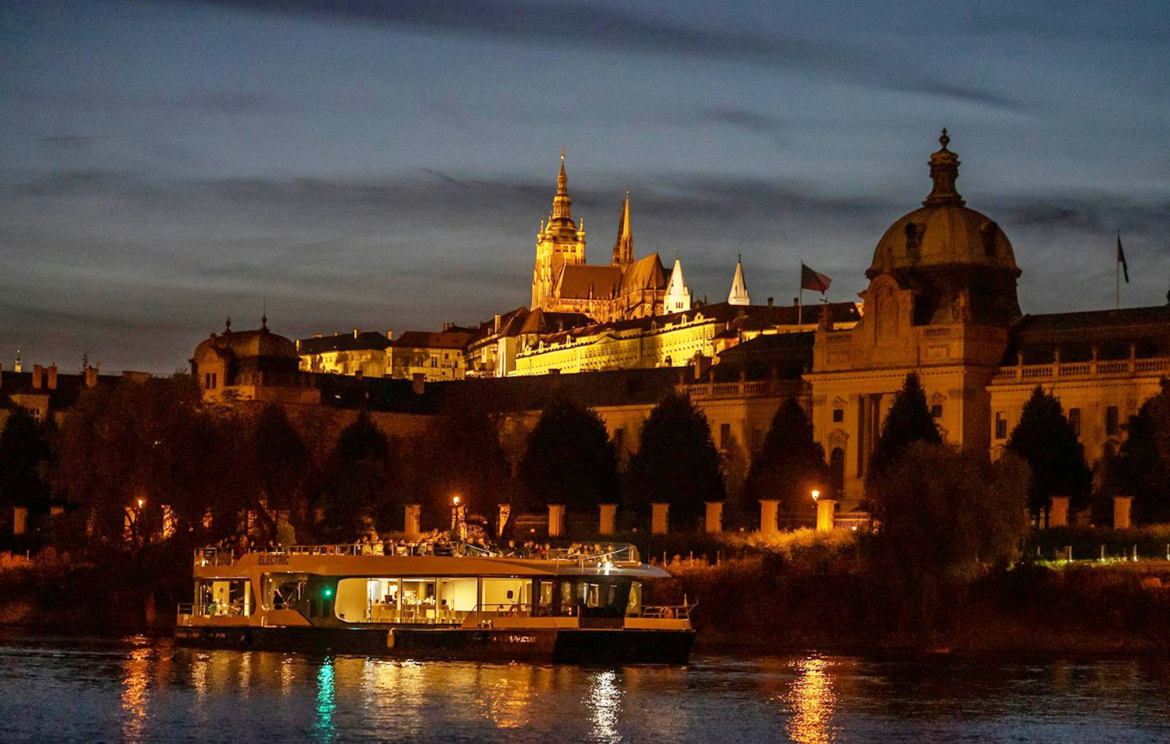 Prague river cruise boat at night with illuminated castle in the background.