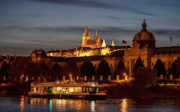 Prague river cruise boat at night with illuminated castle in the background.