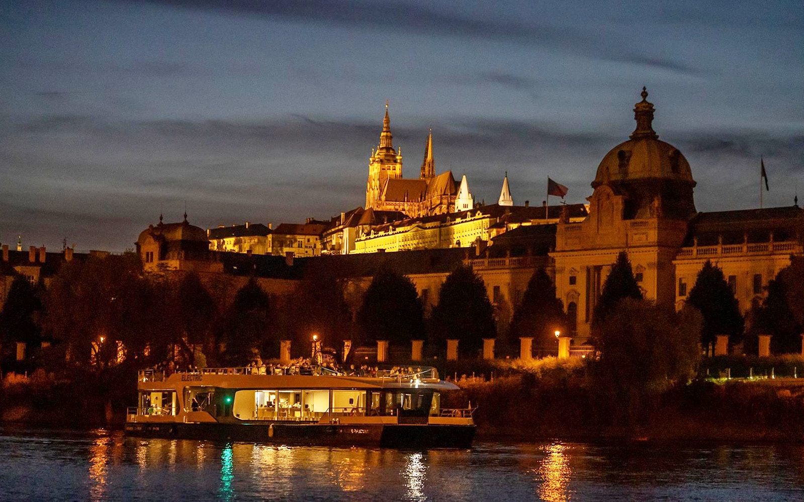 Prague river cruise boat at night with illuminated castle in the background.