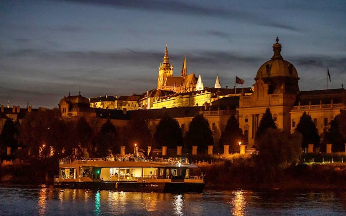 Prague river cruise boat at night with illuminated castle in the background.