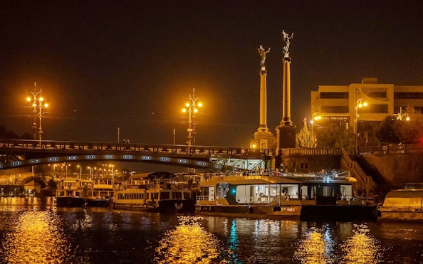 Prague river cruise boat at night near illuminated bridge and statues.