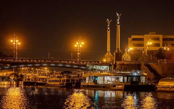 Prague river cruise boat at night near illuminated bridge and statues.