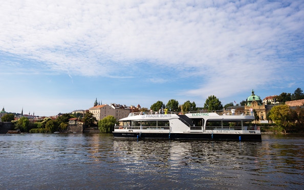 Electric boat cruising on the Vltava River in Prague with historic buildings in the background.