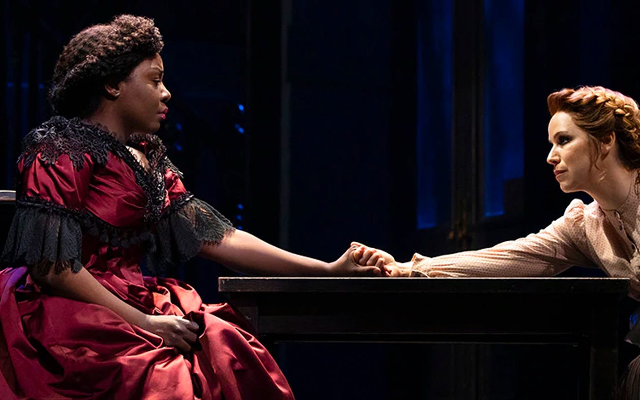 Two actors in a dramatic scene from Paradise Square, holding hands across a table.