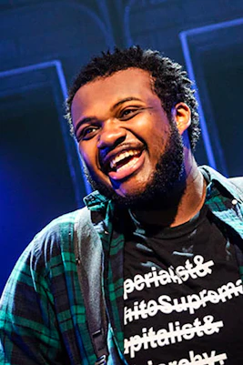 Performer on stage during "A Strange Loop" show, wearing a plaid shirt and graphic tee.