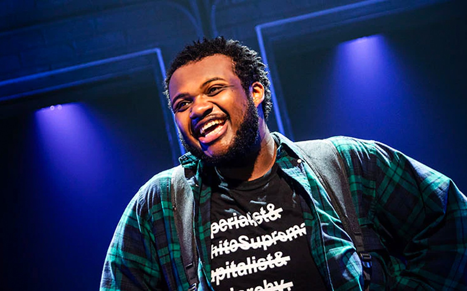Performer on stage during "A Strange Loop" show, wearing a plaid shirt and graphic tee.