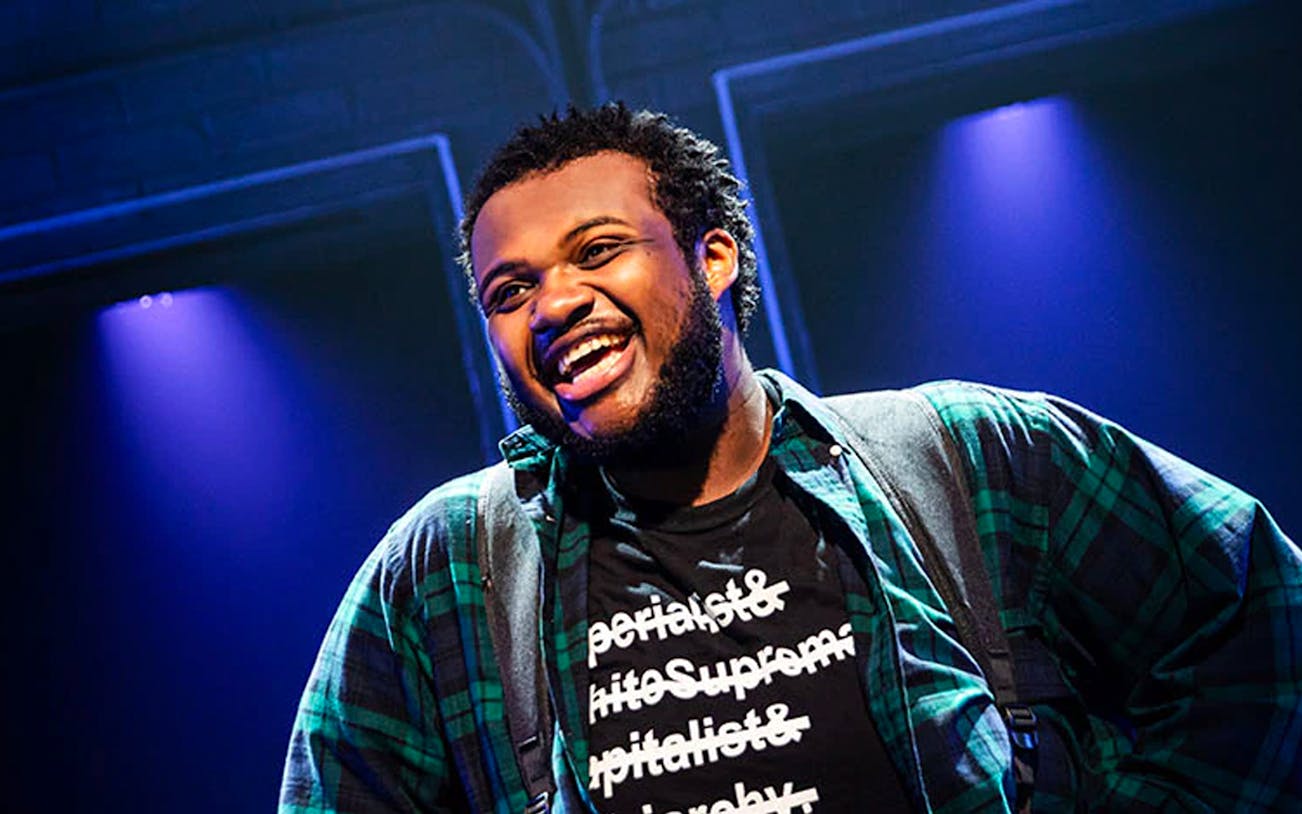 Performer on stage during "A Strange Loop" show, wearing a plaid shirt and graphic tee.