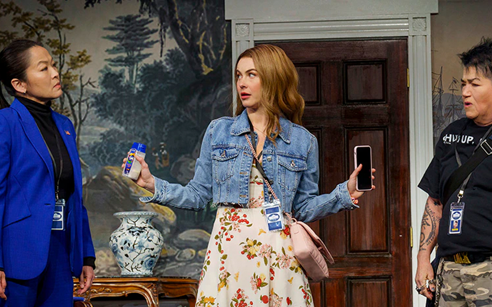 Three women in conversation, one holding a phone and a bottle, in a room with a decorative vase.