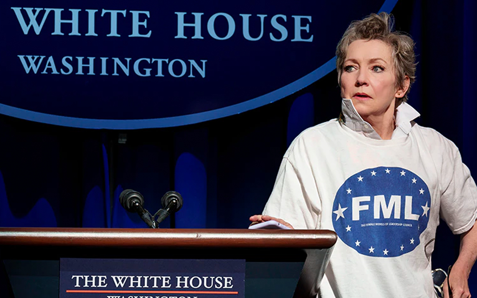 A person stands at a podium with a White House backdrop, wearing a shirt with "FML" logo.