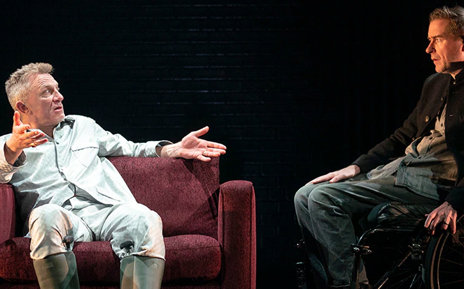 Two actors in a dramatic scene from Macbeth, one seated on a couch, the other in a wheelchair.