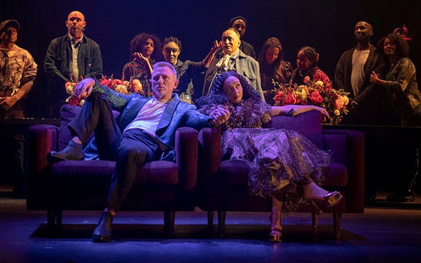 Actors in a modern stage production of Macbeth, seated on a couch with a cast ensemble in the background.