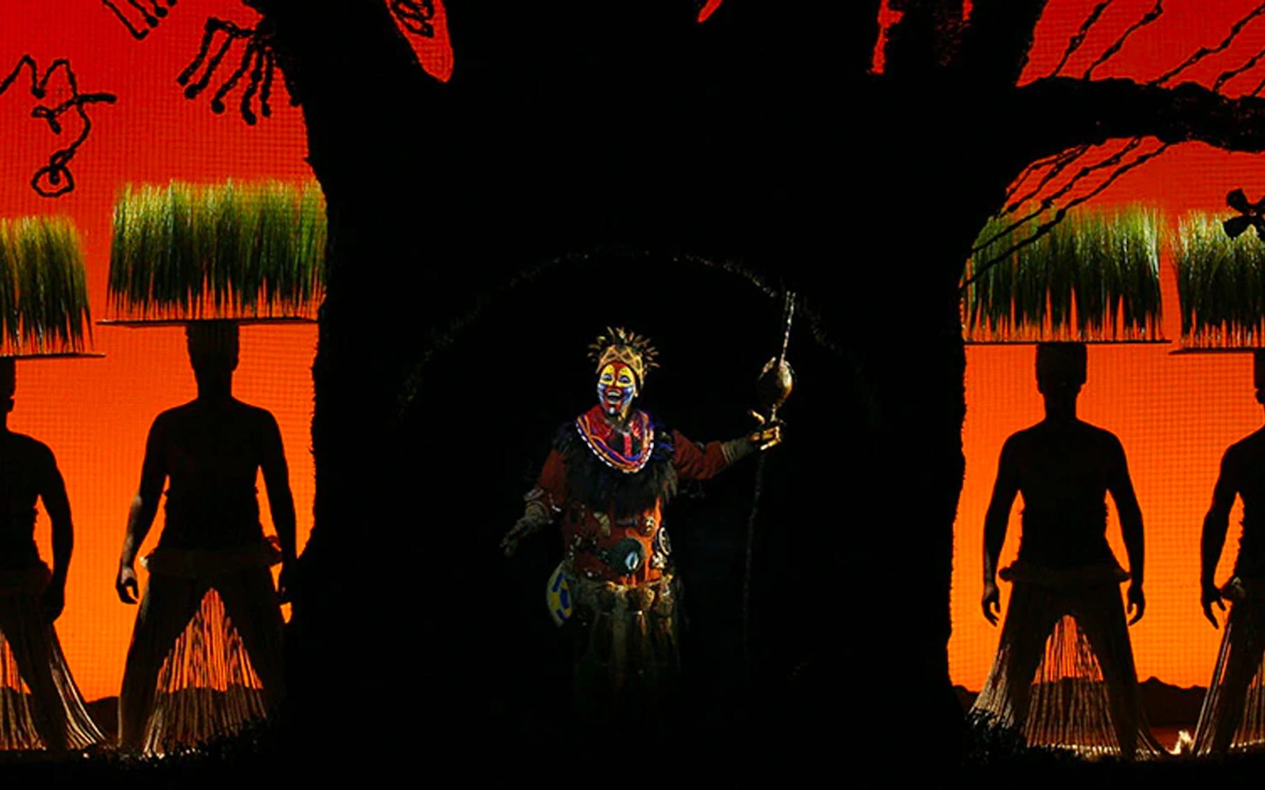 Rafiki performing in The Lion King musical with vibrant sunset backdrop.