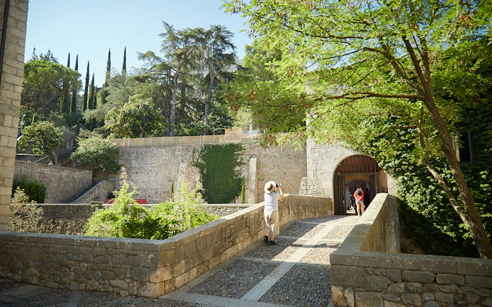 Stone bridge leading to historic entrance in Girona, surrounded by lush greenery.