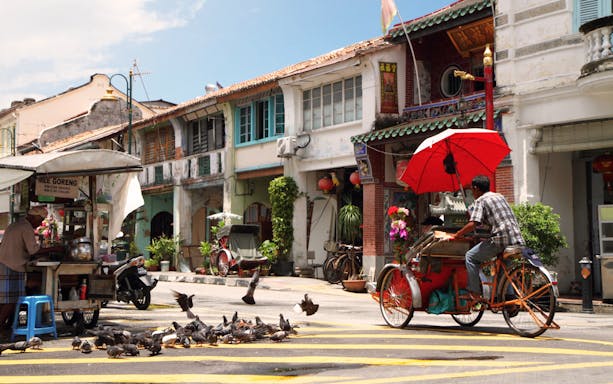 Trishaw rider in Georgetown, Penang, passing heritage shophouses and street food vendor.