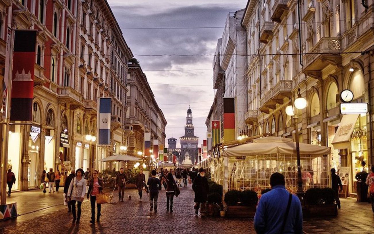 Street view of Milan with people walking and the Sforza Castle in the background.