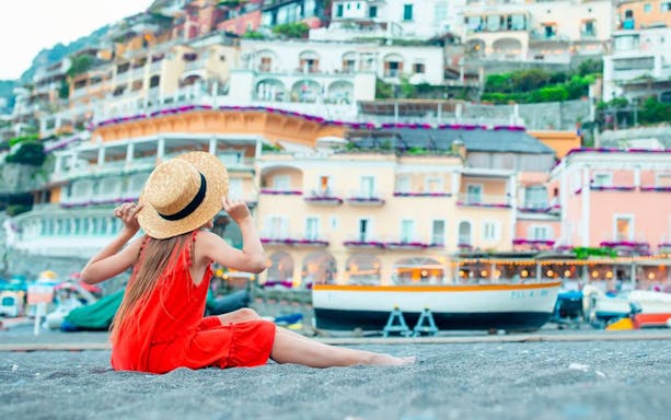 Person in a red dress sitting on Positano beach, colorful cliffside buildings in view.