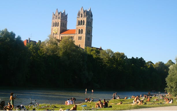 People relaxing by the river with bicycles near a historic church in the English Garden, Munich.