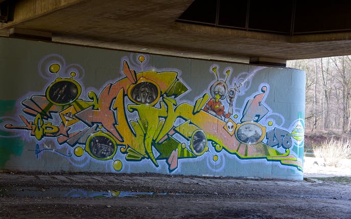 Street art mural featuring colorful abstract shapes and alien figures under a bridge in Munich.