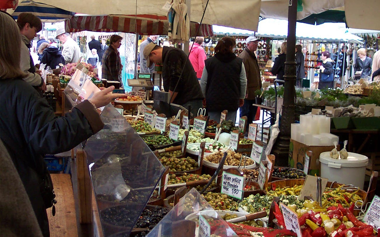 Bavarian market with people browsing stalls of olives, peppers, and local produce.