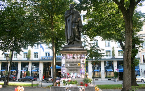 Statue in Munich park with flowers and photos at the base, near Bayerischer Hof hotel.