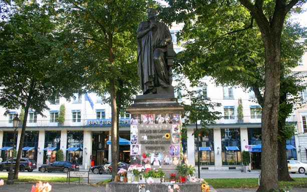 Statue in Munich park with flowers and photos at the base, near Bayerischer Hof hotel.