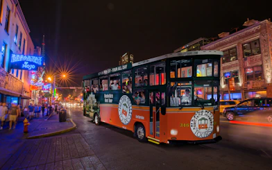 Hop-on hop-off trolley on a lively Nashville street at night, passing neon-lit bars and restaurants.