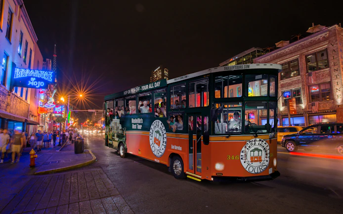 Hop-on hop-off trolley on a lively Nashville street at night, passing neon-lit bars and restaurants.