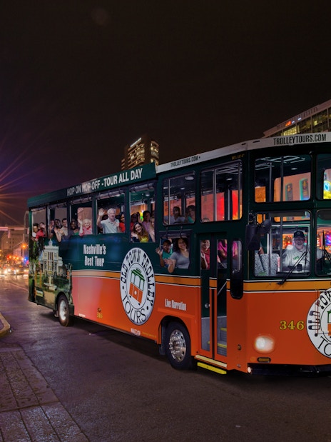 Hop-on hop-off trolley on a lively Nashville street at night, passing neon-lit bars and restaurants.