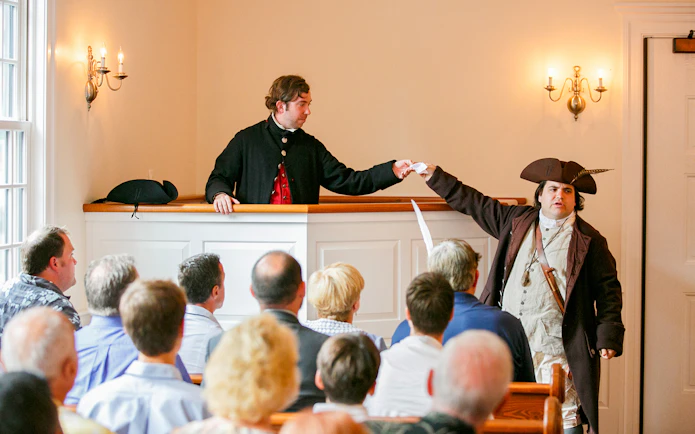 Reenactment of Boston Tea Party debate with actors in period costumes inside a historic room.