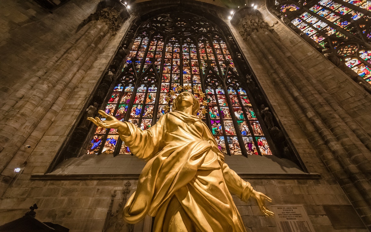 Golden statue inside Milan Duomo with stained glass windows in the background.