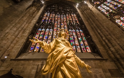 Golden statue inside Milan Duomo with stained glass windows in the background.