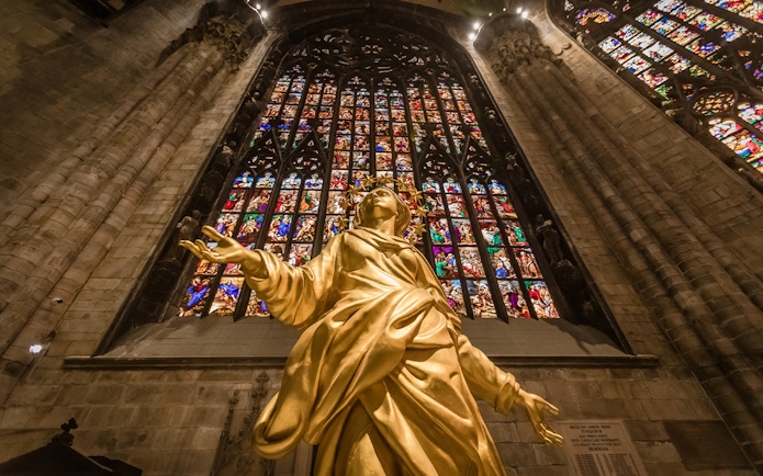 Golden statue inside Milan Duomo with stained glass windows in the background.