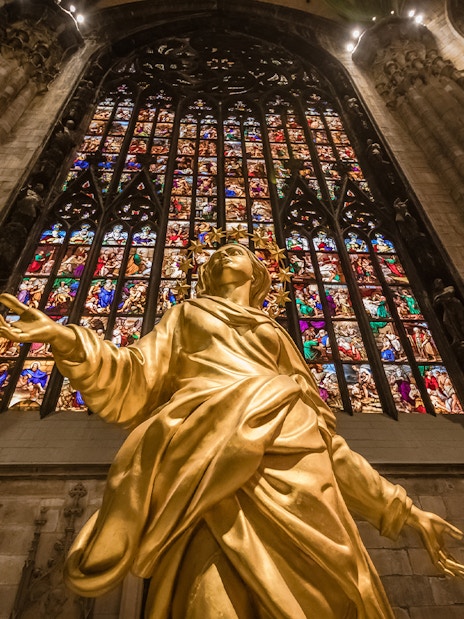 Golden statue inside Milan Duomo with stained glass windows in the background.