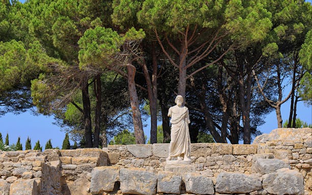 Statue among ancient stone ruins at Empúries, surrounded by trees.