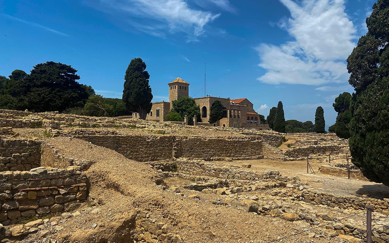 Ancient stone ruins of Empúries with a historic building and trees in the background, Catalonia, Spain.