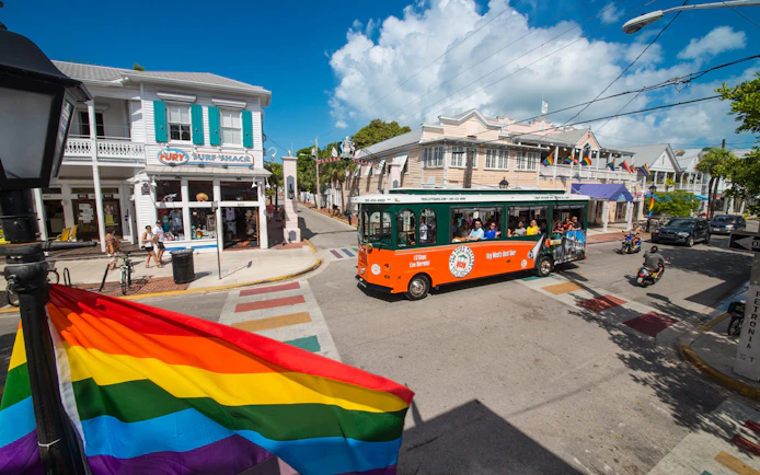 Key West Old Town Trolley passing colorful shops and rainbow flag on a sunny street.